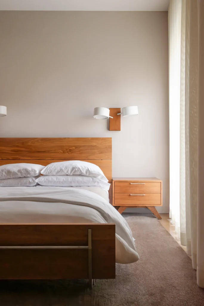Minimalist bedroom with a wooden bed frame, white bedding, matching nightstand, and soft natural light.