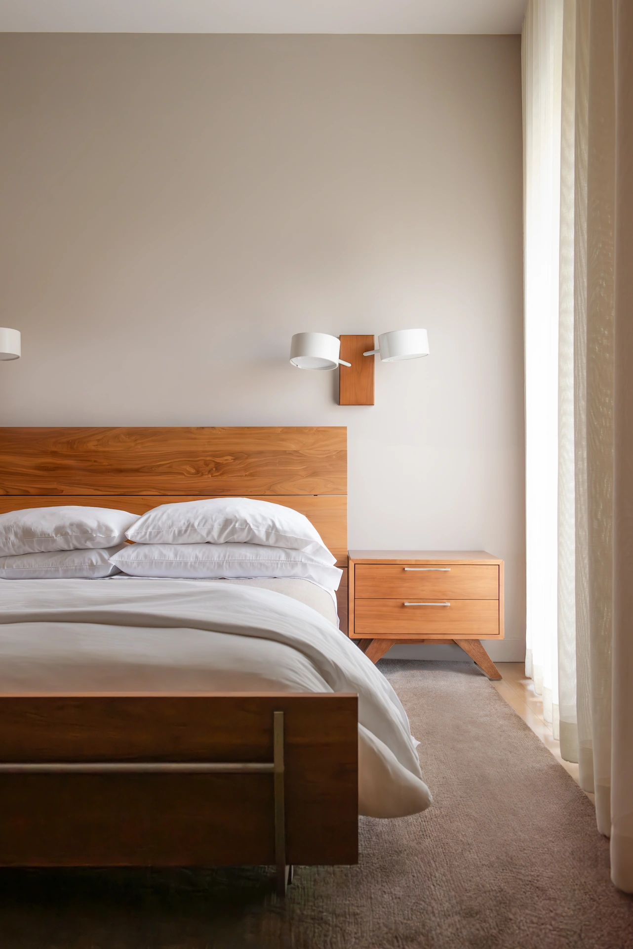Minimalist bedroom with a wooden bed frame, white bedding, matching nightstand, and soft natural light.