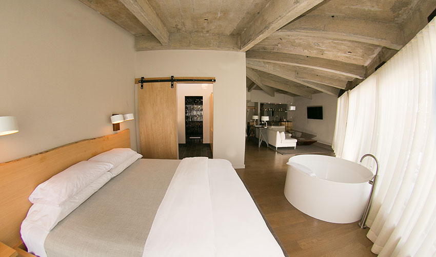 Open-concept suite with exposed concrete ceiling, wooden bed, freestanding soaking tub, and floor-to-ceiling curtains.