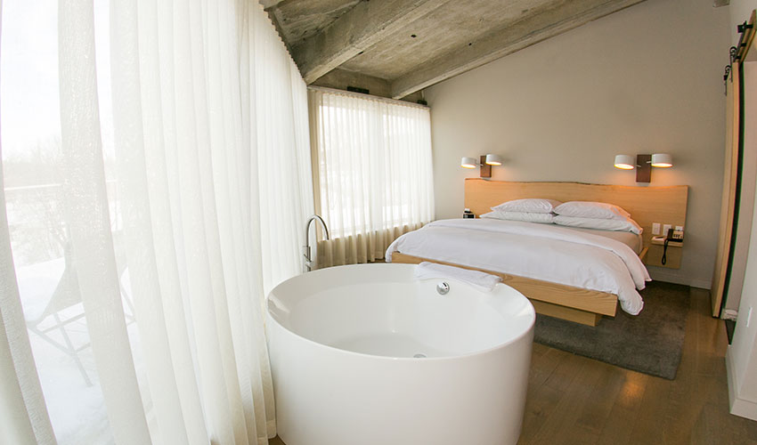 Modern bedroom with a freestanding soaking tub by sheer curtains, a low platform bed, and minimalist lighting.