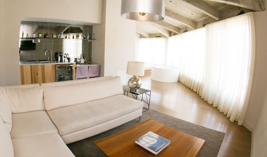 Bright modern living suite with a sofa, kitchenette, wood floors, large windows, and sheer curtains letting in natural light.