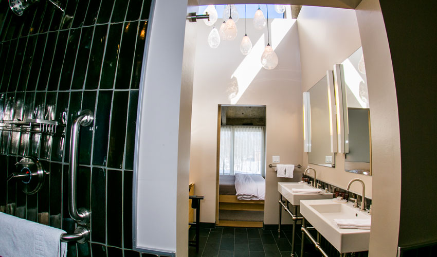 Modern bathroom with black tile shower, double sinks, pendant lights, and a view into a bedroom beyond.