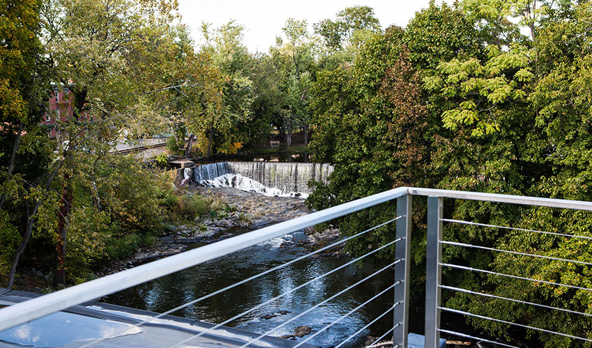 View from a balcony overlooking a river and small waterfall, surrounded by lush green trees.