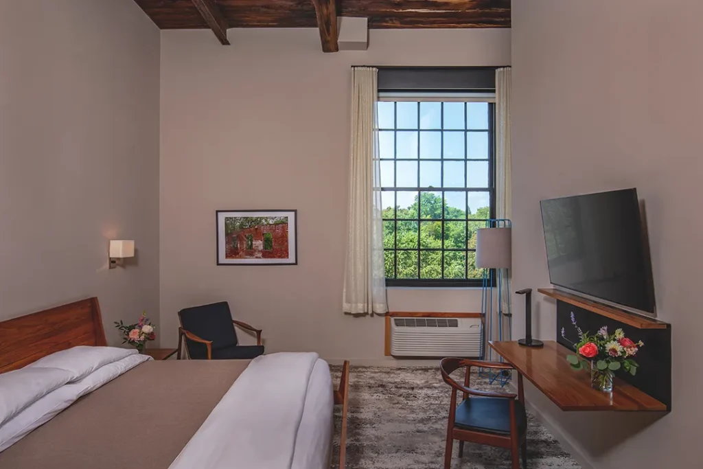 Modern hotel room with exposed beams, large window, bed, desk, and seating area.