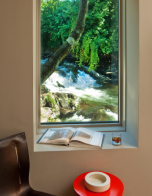 Open book and coffee on a red table beside a window overlooking a flowing stream and green trees.