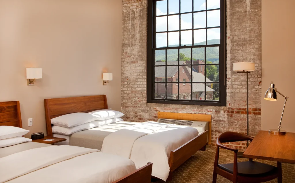 Bright hotel room with two beds, exposed brick walls, wood furnishings, and a large window overlooking rooftops and hills.