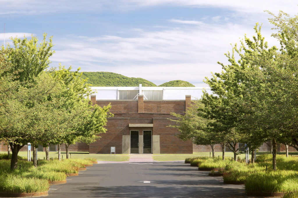 Brick building framed by trees at the end of a landscaped pathway, with green hills and a bright sky in the background.