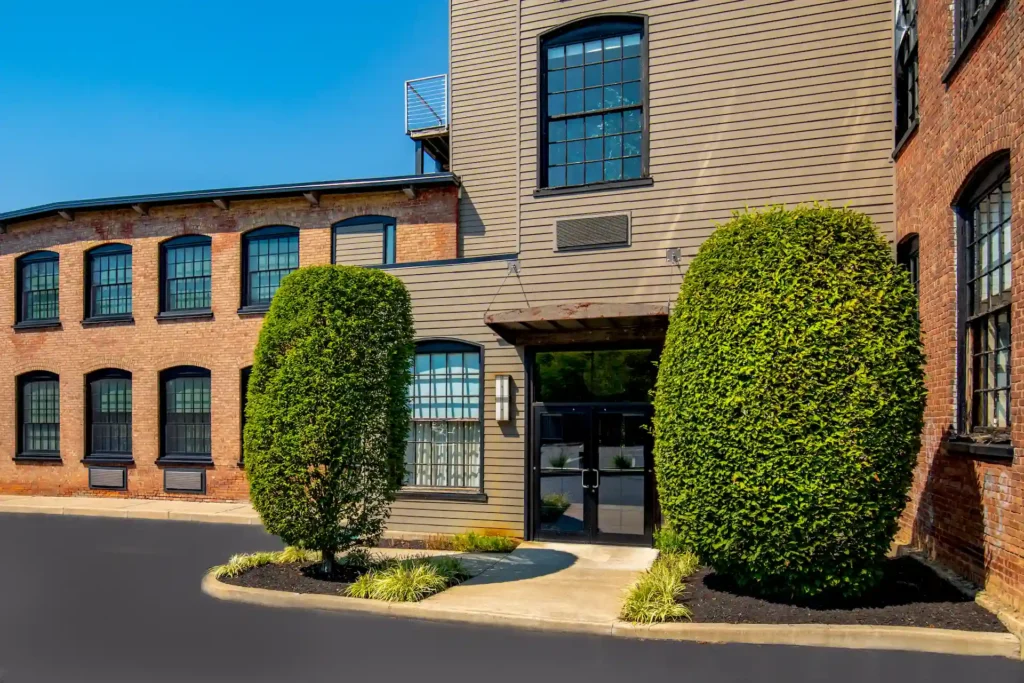 Building entrance with glass doors, wood siding, and brick walls, framed by trimmed hedges under a clear blue sky.