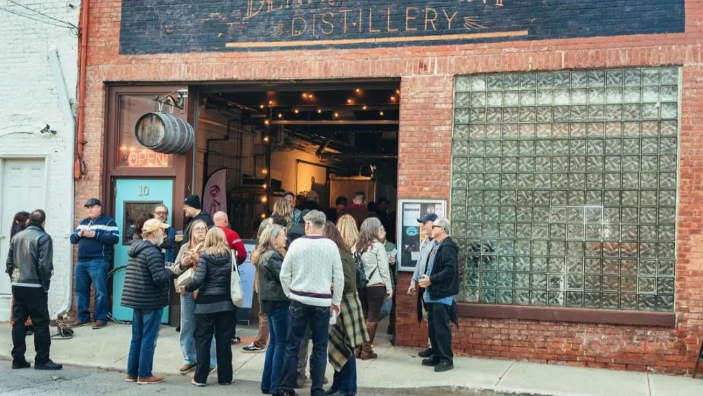 Crowd gathers outside a brick distillery entrance with open doors and string lights.