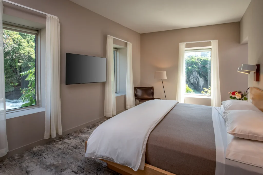 Modern bedroom with a neatly made bed, large windows, soft neutral tones, and natural light with greenery visible outside.