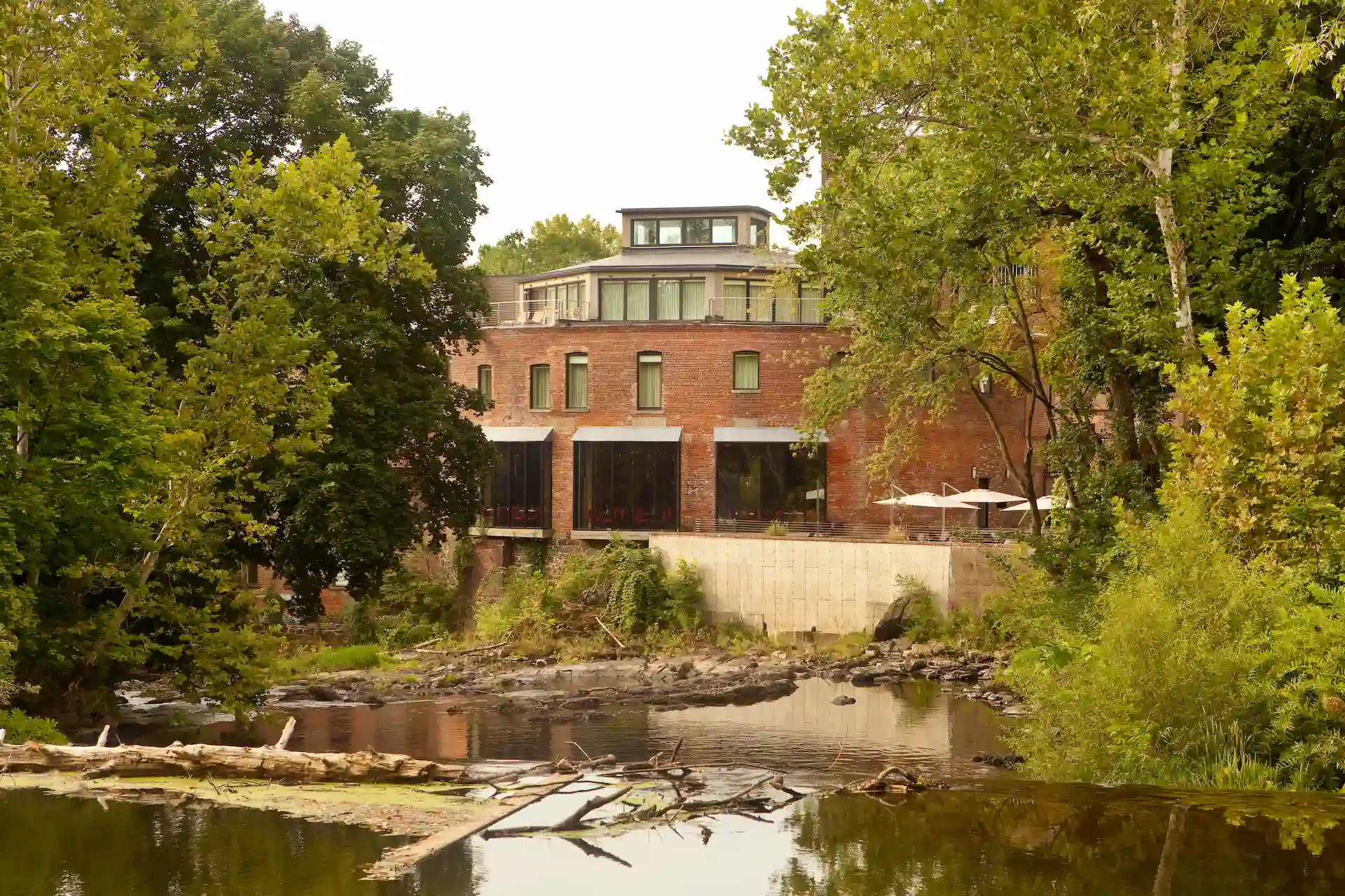 Brick building set along a calm river, surrounded by trees and greenery with outdoor seating visible on a terrace.
