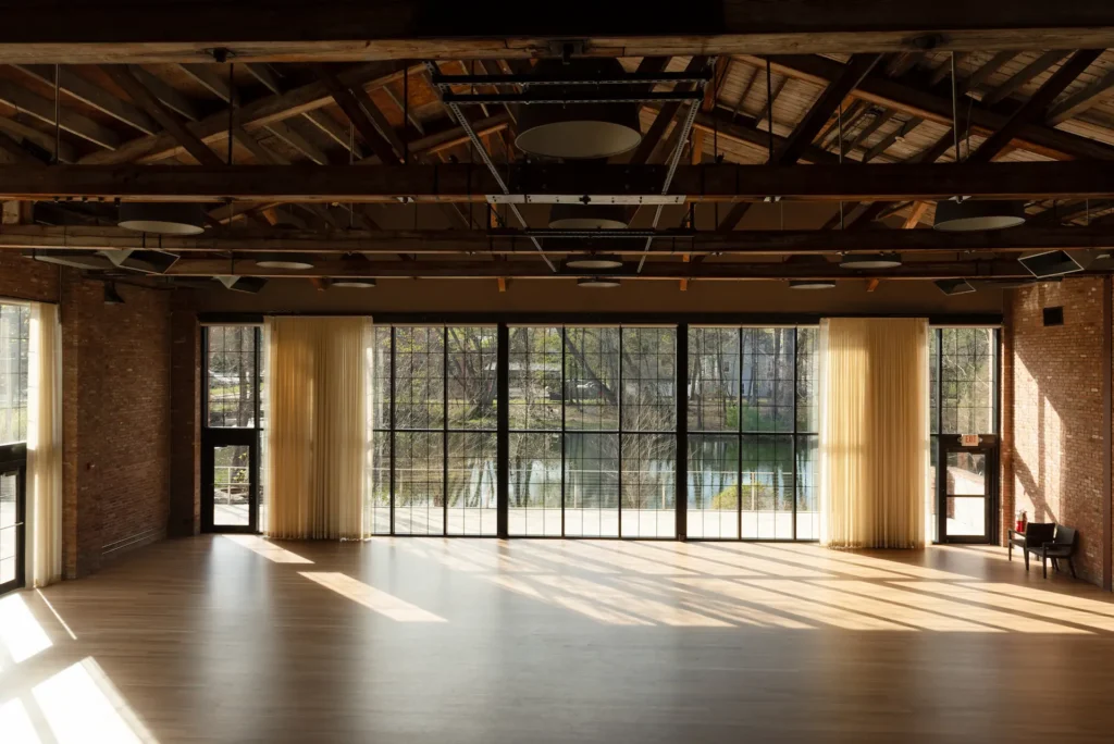 Sunlit empty industrial event hall with wood floors, exposed beams, and large windows overlooking trees.