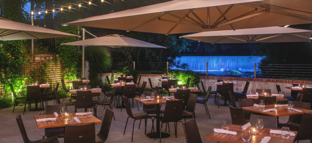 Outdoor dining patio at night with candlelit tables, umbrellas, string lights, and a softly lit waterfall backdrop.