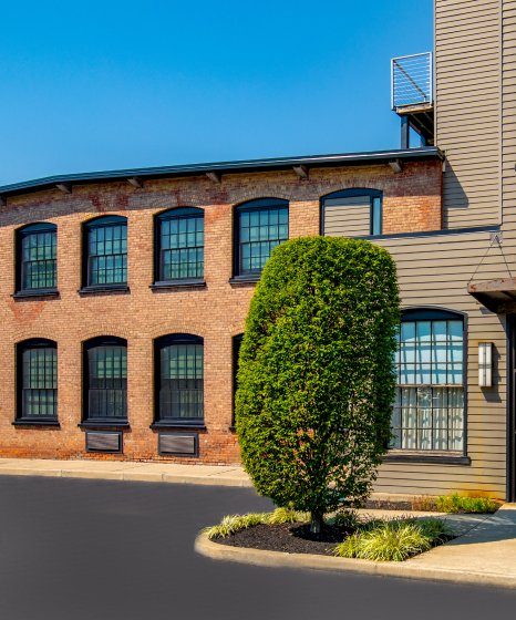 Exterior of a brick and modern-sided building with large windows, a manicured tree, and a clear blue sky.