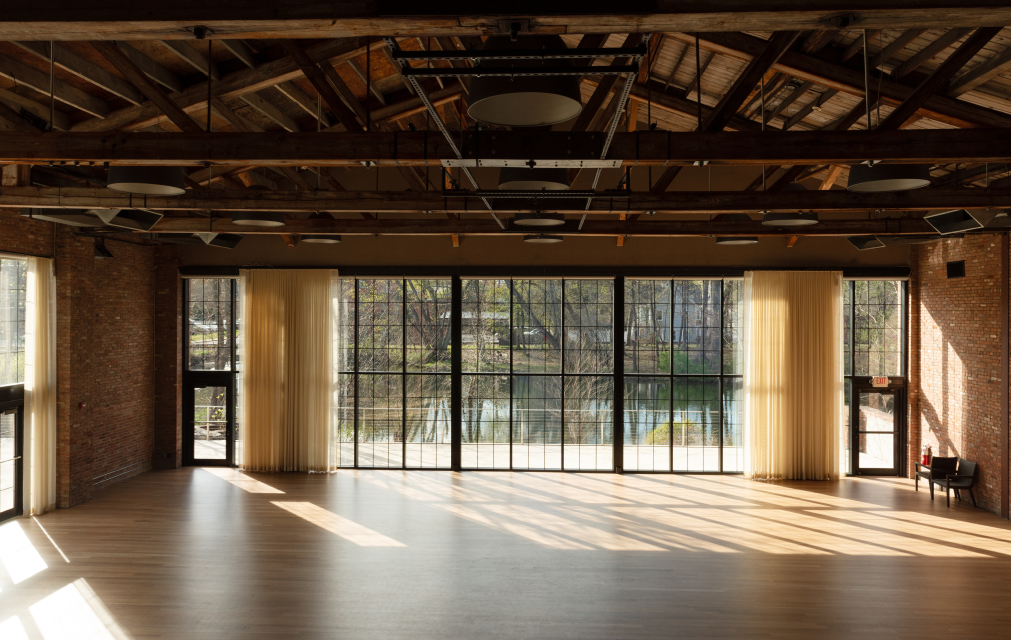 Sunlit industrial event space with exposed beams, brick walls, wood floors, and large windows overlooking water.