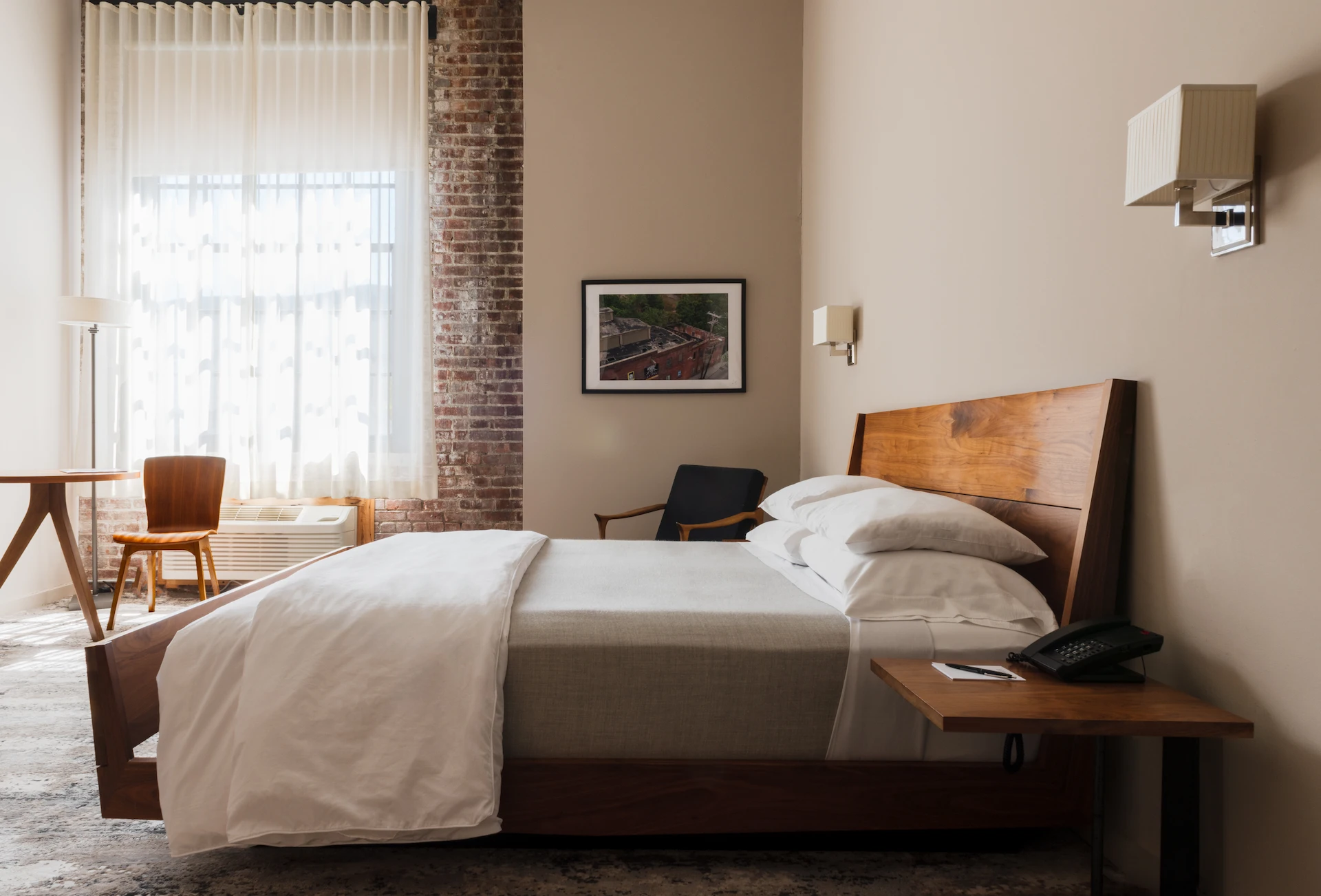 Modern guest room with a wooden bed, white linens, exposed brick wall, desk seating, and soft daylight.