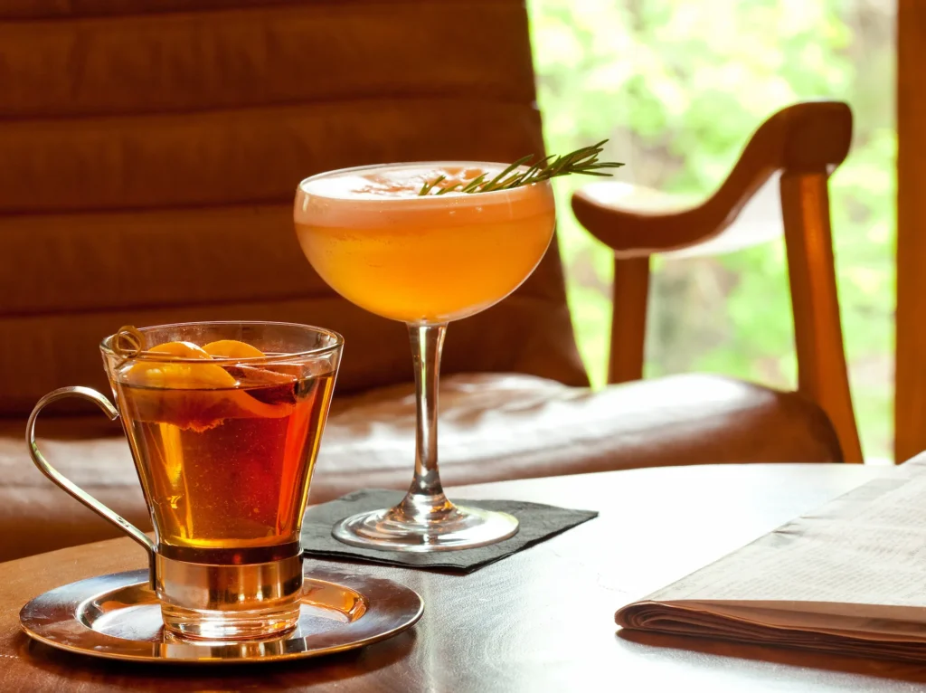Cocktail and hot tea on a table by a window, with a newspaper nearby and warm seating in the background.