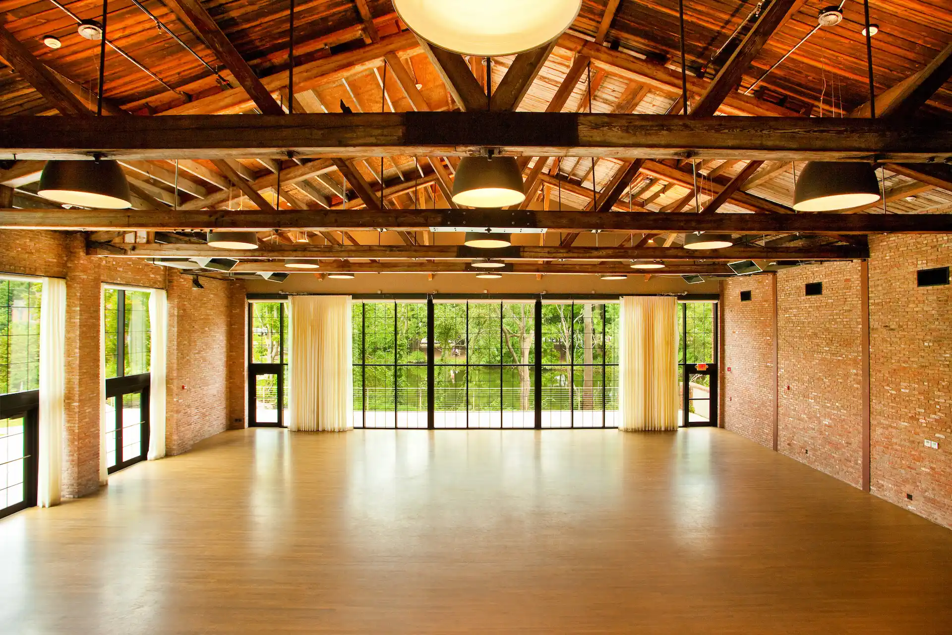 Spacious empty event hall with exposed wooden beams, brick walls, and large windows overlooking greenery.