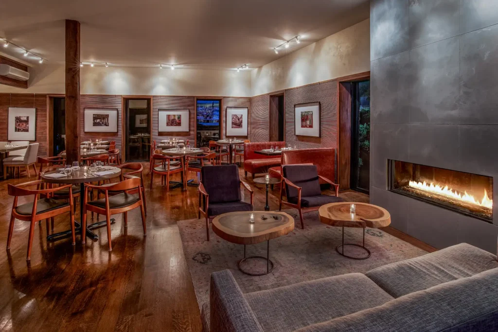 Warm, modern restaurant interior with wood floors, set tables, lounge seating, and a glowing fireplace along the wall.