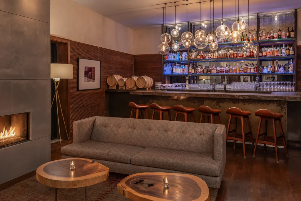 Cozy bar lounge with stocked backlit shelves, hanging globe lights, bar stools, and a fireplace seating area.