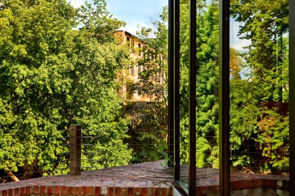 View through large windows onto a brick terrace and lush green trees, with a historic building partially visible beyond.