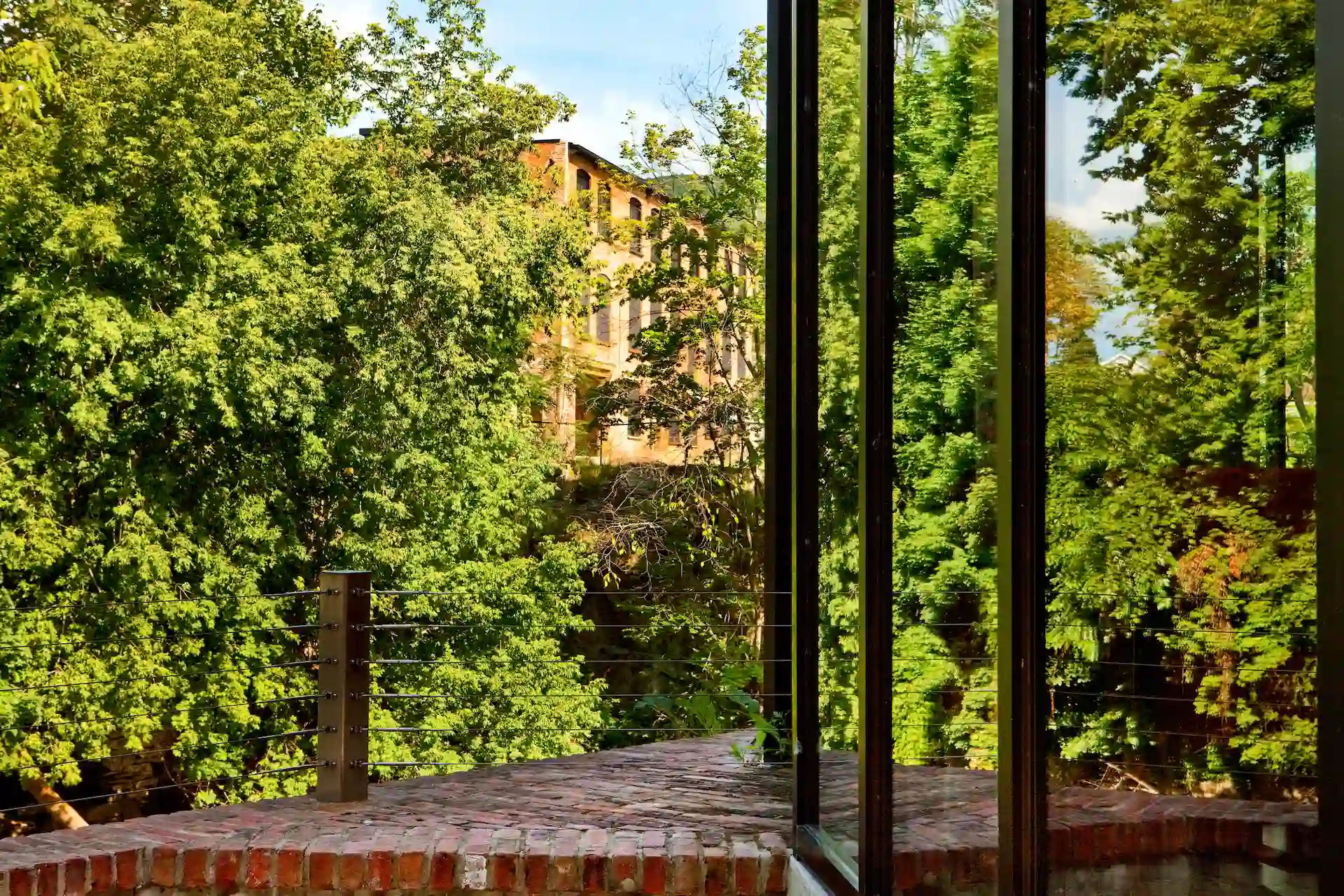 View through large windows onto a brick terrace and lush green trees, with a historic building partially visible beyond.