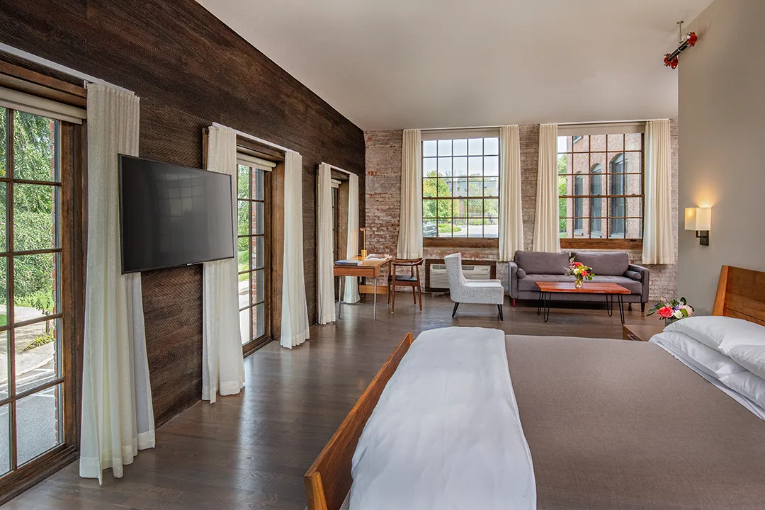 Spacious modern hotel room with wooden bed, seating area, large windows, and mounted tv.