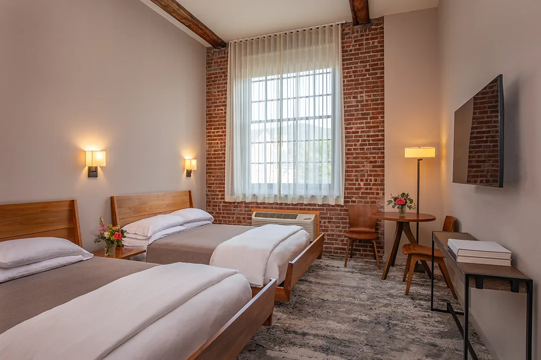 Twin-bed hotel room with brick walls, wooden beds, desk, and large window.