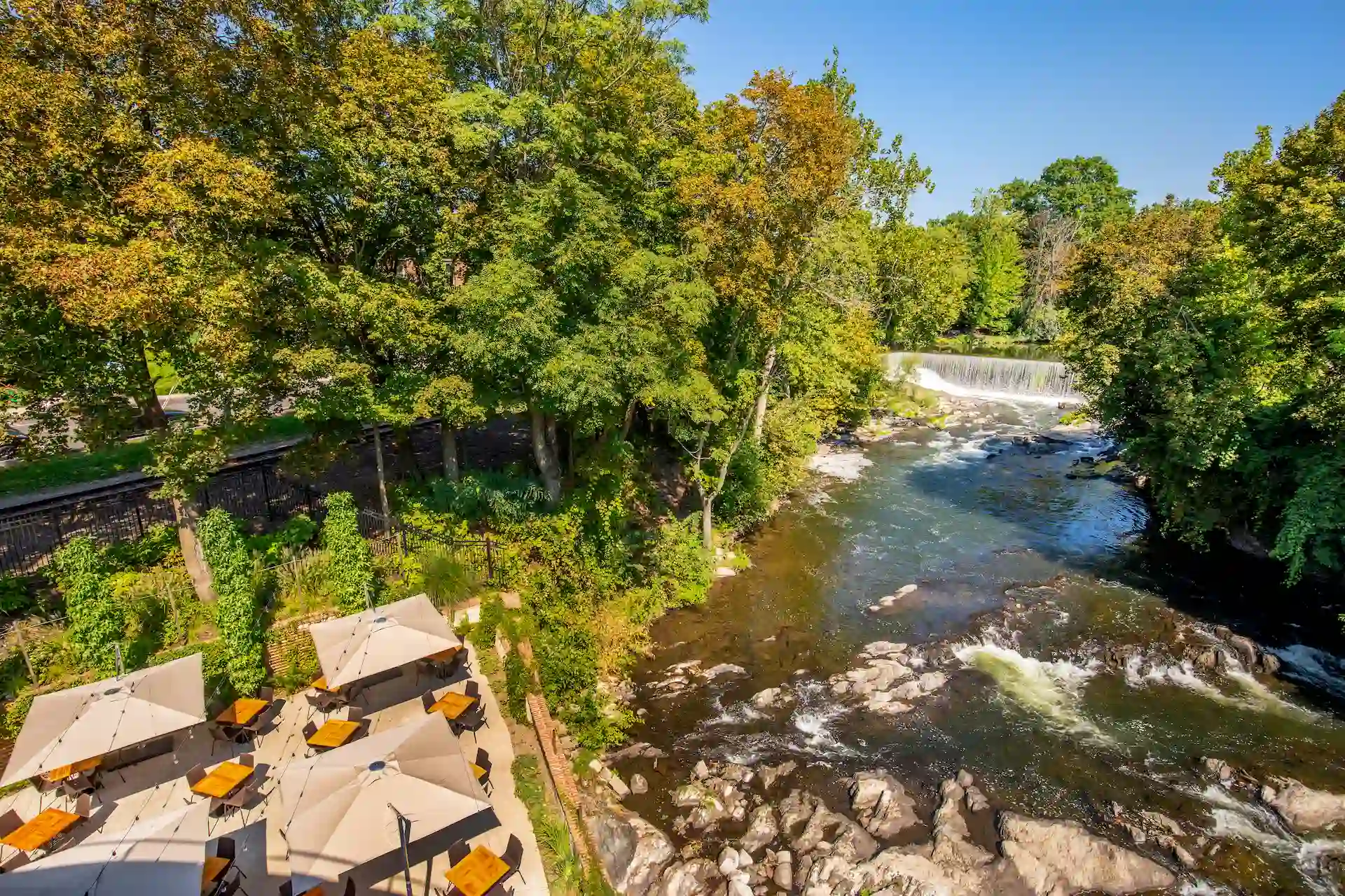 Aerial view of outdoor dining patios beside a flowing river and waterfall, surrounded by trees and greenery.