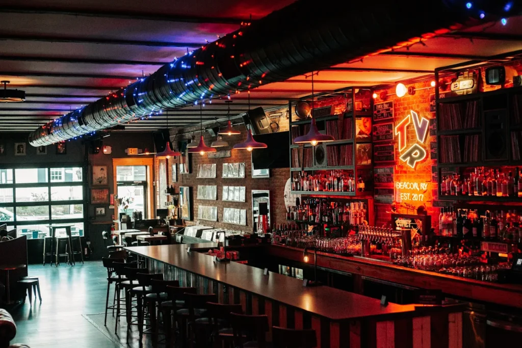 Moody bar interior with long counter, shelves of bottles, neon sign, and colorful string lights.