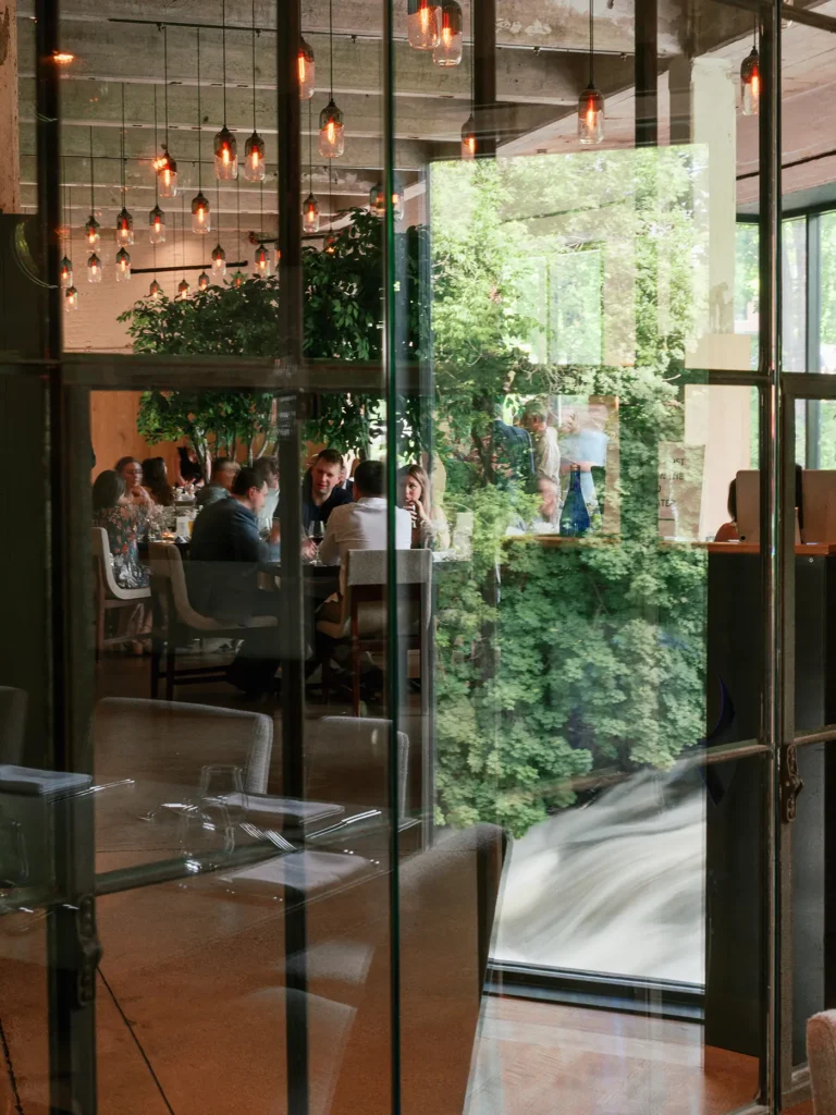 Guests dine and converse inside a modern restaurant, seen through glass with hanging lights and greenery reflected outside.