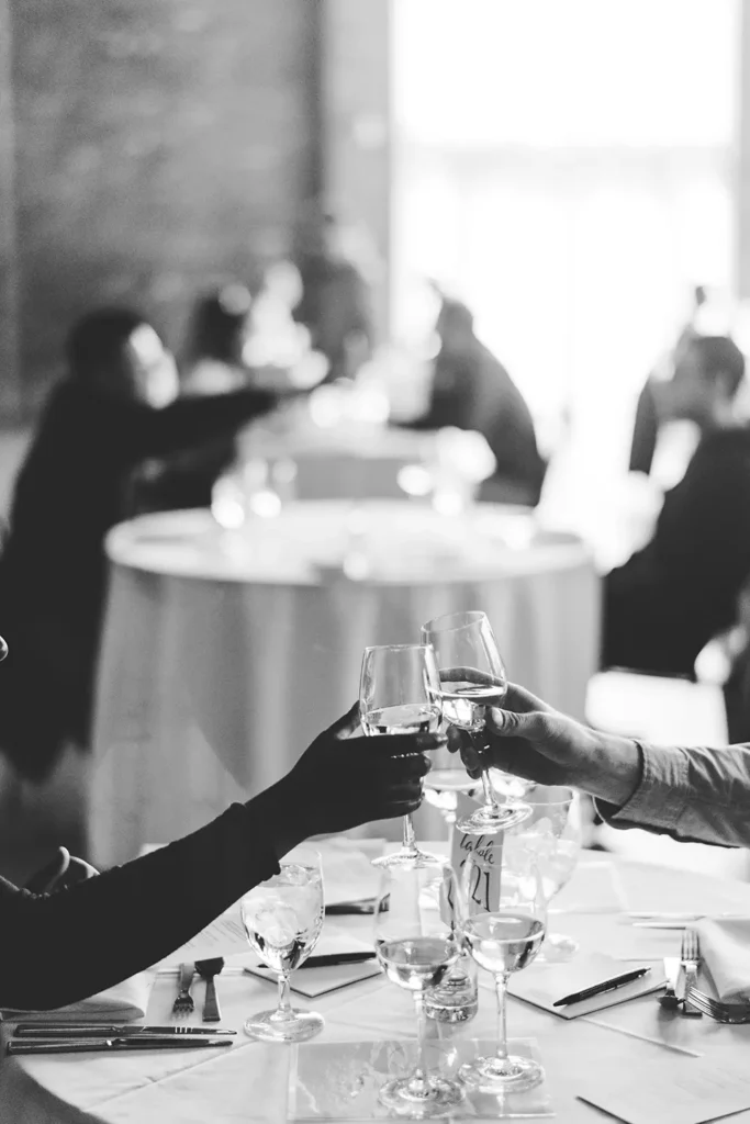 Two guests clink wine glasses in a toast at a formal event, seated at a round table with glassware and place settings.