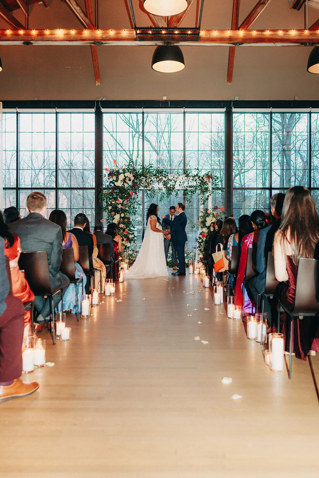 Indoor wedding ceremony with couple exchanging vows beneath a floral arch and candlelit aisle.