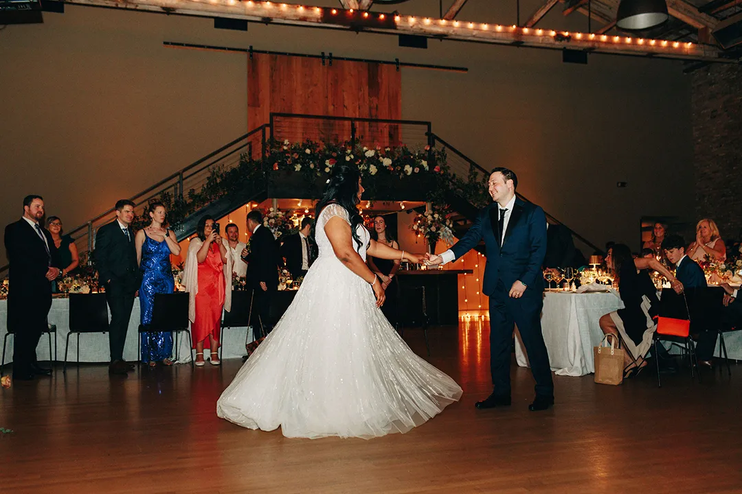 Bride and groom holding hands during their first dance as guests watch in a warmly lit venue.