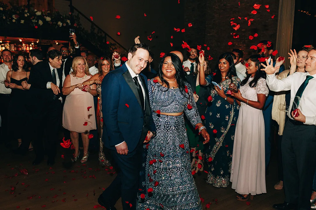 Couple walk through cheering guests as rose petals fall at a wedding celebration.