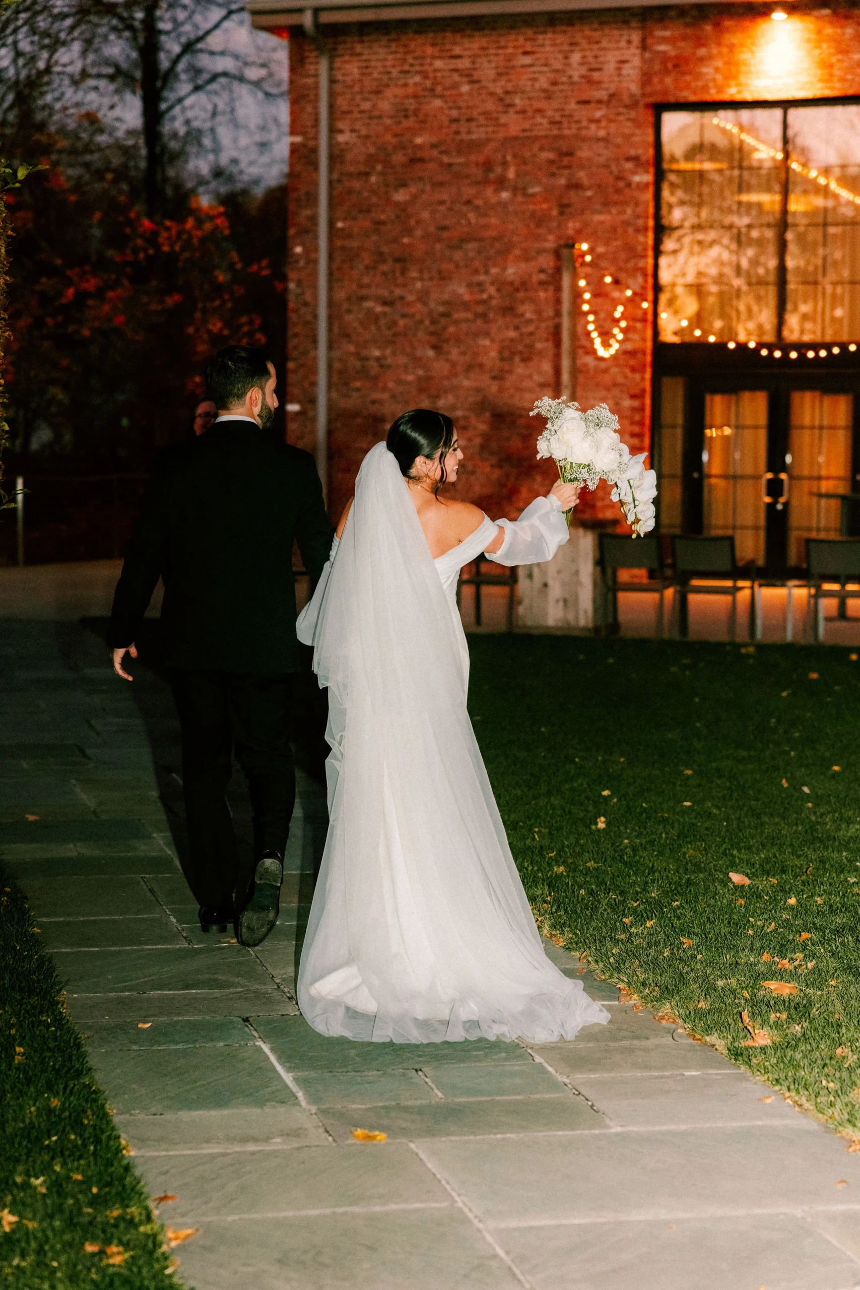 Bride and groom walk toward a brick venue at dusk, bride lifting bouquet as warm string lights glow inside.