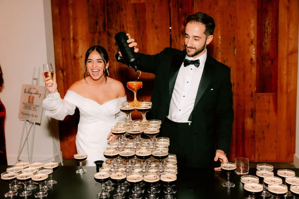 Bride and groom pour champagne into a glass tower, laughing during their wedding celebration.