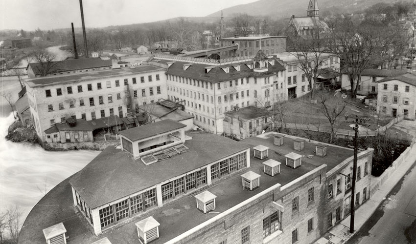 Aerial view of historic factory buildings along a river, with multiple brick structures, smokestacks, and surrounding hills.