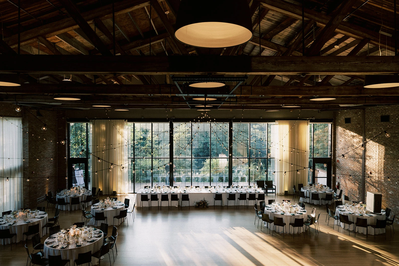 Elegant event hall with round tables, string lights, exposed wood beams, and floor-to-ceiling windows overlooking greenery.