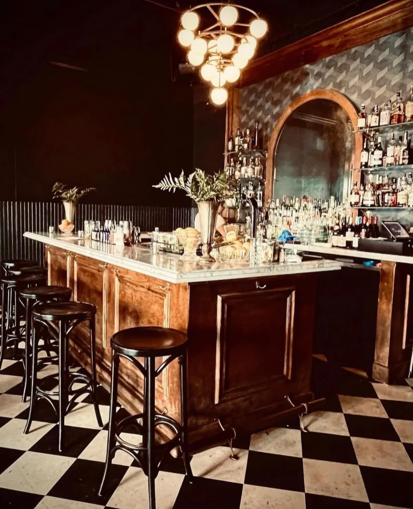 Vintage-style bar with marble counter, stools, liquor shelves, and checkered floor.