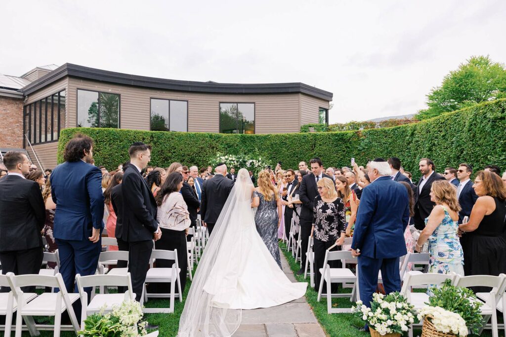 Bride walks down an outdoor aisle toward the ceremony, surrounded by standing guests and greenery at a garden wedding.
