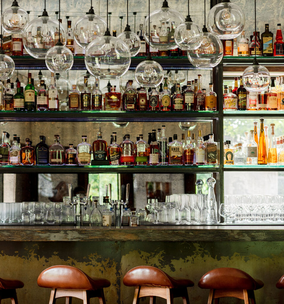 Backlit bar shelves lined with liquor bottles, glassware, and globe pendant lights above leather bar stools.