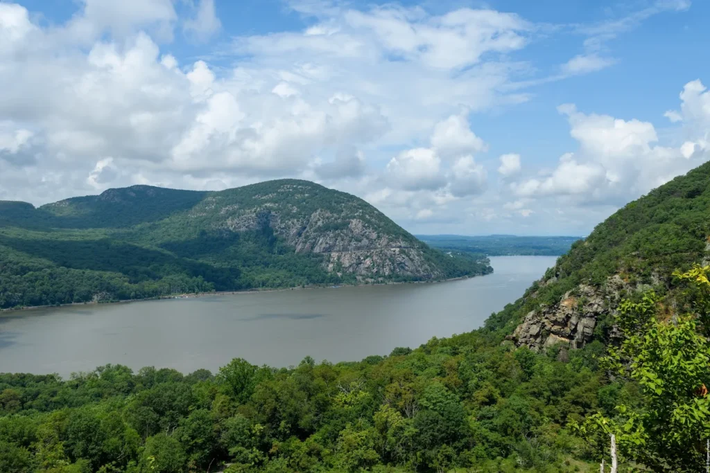 Wide river winding between forested hills under a bright, cloud-filled sky.
