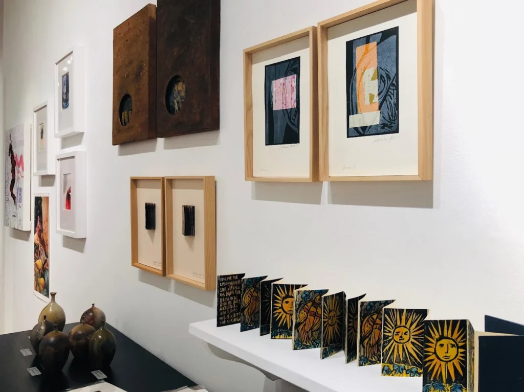 Gallery wall with framed mixed-media artworks and small illustrated prints on a display shelf.