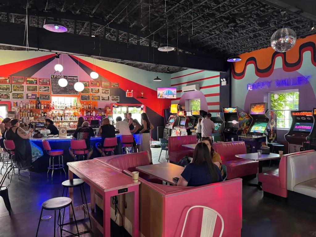 Colorful retro arcade bar with pink booths, game cabinets, neon lights, and patrons socializing.