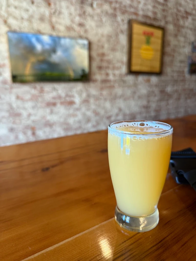 Glass of hazy pale beer on a wooden bar with brick wall and framed art behind.
