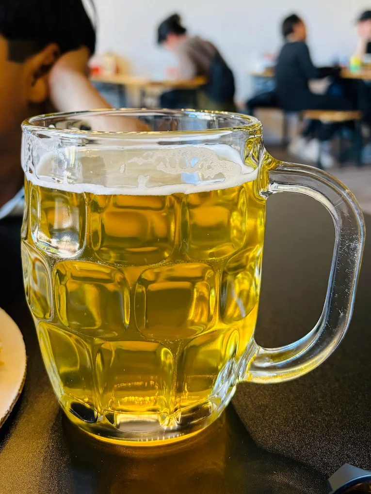 Clear glass mug of golden beer with foam on a table, café patrons blurred in background.