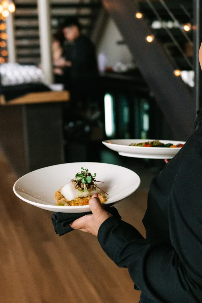 Server carrying plated gourmet dishes in a softly lit indoor venue with bar in background.