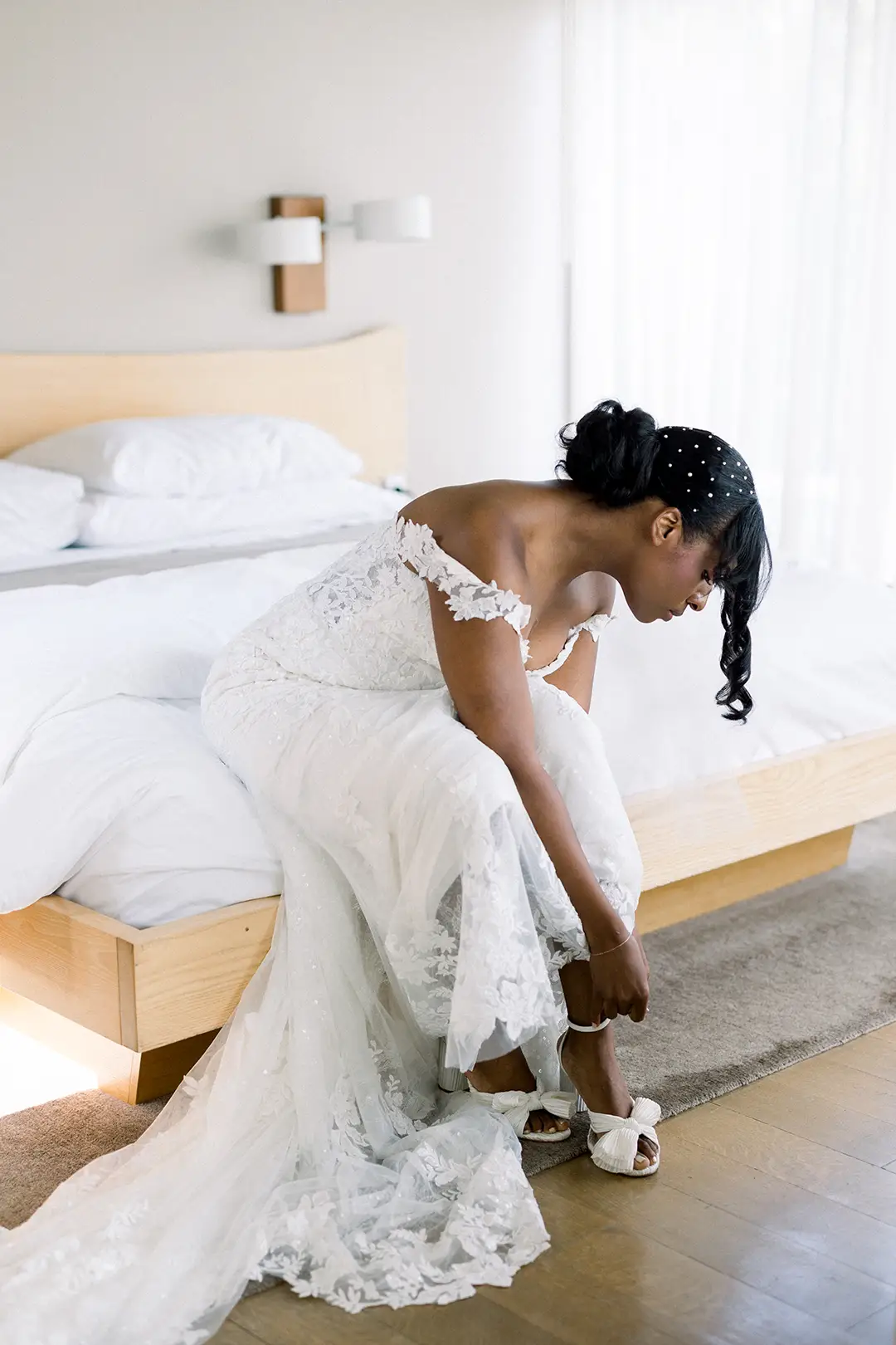 Bride sitting on a bed adjusting her shoe in a bright, minimalist hotel room.