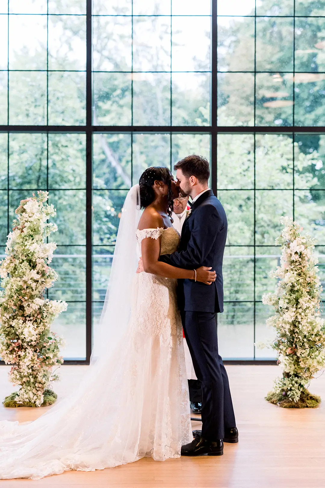 Bride and groom kiss during their ceremony framed by flowers and large windows.